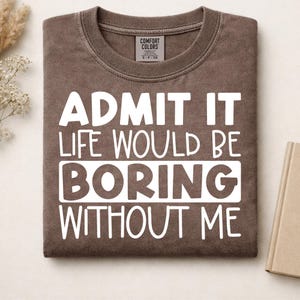 May include: A brown crewneck t-shirt with white text that reads: ADMIT IT LIFE WOULD BE BORING WITHOUT ME. The shirt features a Comfort Colors brand label at the inner neckline and has a soft, washed-out appearance with a ribbed collar.