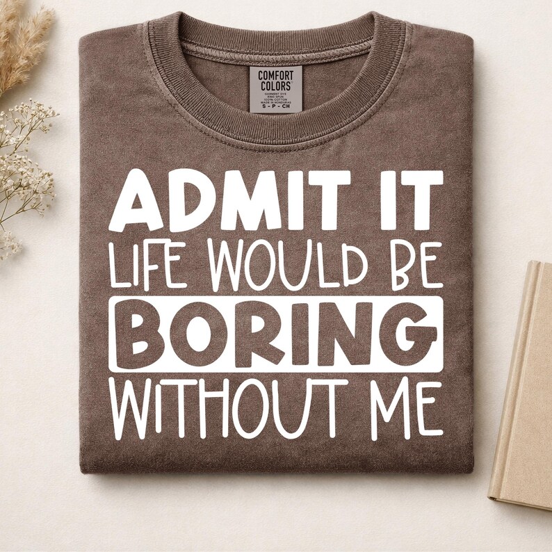 May include: A brown crewneck t-shirt with white text that reads: ADMIT IT LIFE WOULD BE BORING WITHOUT ME. The shirt features a Comfort Colors brand label at the inner neckline and has a soft, washed-out appearance with a ribbed collar.