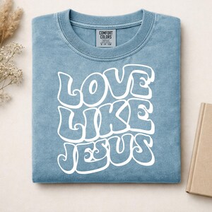 Pu&ograve; includere: Una t-shirt blu a maniche corte con scollo rotondo e un'etichetta del marchio Comfort Colors all'interno del collo. La parte anteriore presenta una scritta bianca in un carattere ondulato e stilizzato che recita LOVE LIKE JESUS, centrata sul petto.