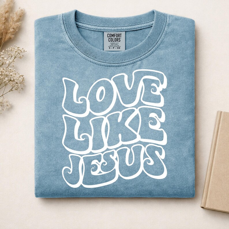 Pu&ograve; includere: Una t-shirt blu a maniche corte con scollo rotondo e un'etichetta del marchio Comfort Colors all'interno del collo. La parte anteriore presenta una scritta bianca in un carattere ondulato e stilizzato che recita LOVE LIKE JESUS, centrata sul petto.