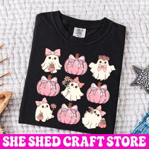 K&ouml;nnte beinhalten: Ein schwarzes T-Shirt mit einem Grafikdruck von vier wei&szlig;en Geistern und vier rosa K&uuml;rbissen, die alle mit rosa Schleifen und kleinen Blumen verziert sind. Die Geister haben einfache Gesichtsz&uuml;ge und die K&uuml;rbisse haben gerippte Texturen. Das Shirt hat ein Comfort Colors Etikett.