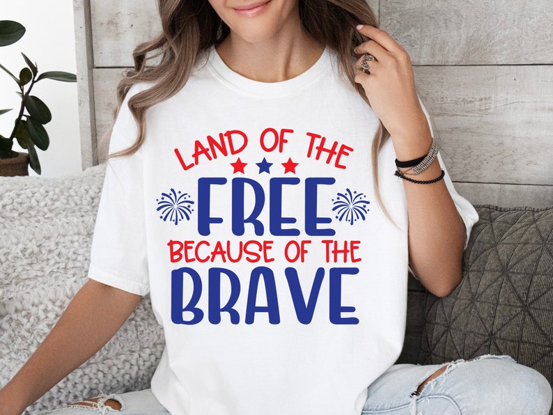 K&ouml;nnte beinhalten: Wei&szlig;es T-Shirt mit einer rot-wei&szlig;-blauen Grafik, auf der "Land of the Free Because of the Brave" steht. Die Grafik enth&auml;lt Sterne und Feuerwerk.