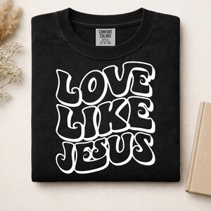 Pu&ograve; includere: Una t-shirt nera a girocollo con la scritta bianca LOVE LIKE JESUS in un carattere audace, ondulato e di ispirazione retr&ograve;. La maglietta presenta un'etichetta del marchio Comfort Colors stampata all'interno del colletto, indicante la taglia S.