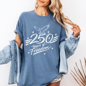 K&ouml;nnte beinhalten: Ein blaues kurz&auml;rmeliges T-Shirt mit einer wei&szlig;en Grafik eines Adlers &uuml;ber dem Text 250 Years of Freedom, umgeben von kleinen Sternen. Das T-Shirt hat einen Rundhalsausschnitt und wird mit einer blauen Jeansjacke &uuml;ber den Schultern getragen.