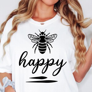 May include: White t-shirt featuring a black graphic of a bee above the word "happy" in a cursive font. Two horizontal black lines are below the word. The shirt is a casual style.