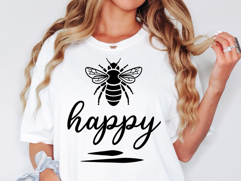 May include: White t-shirt featuring a black graphic of a bee above the word "happy" in a cursive font. Two horizontal black lines are below the word. The shirt is a casual style.