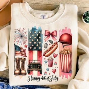 K&ouml;nnte beinhalten: Ein beiger Pullover mit einem grafischen Design, das eine Aquarell-Illustration amerikanischer patriotischer Symbole zeigt, darunter Feuerwerk, eine Flagge, einen Jeep, einen Hotdog, Popcorn, Pommes Frites, eine Schleife und einen Milit&auml;rhelm. Der Text "Happy 4th of July" ist unter dem Grafikdruck.