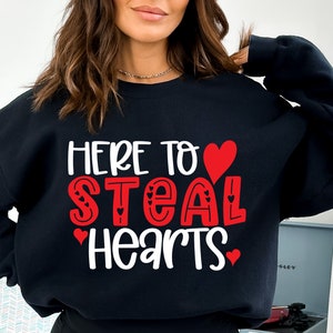 May include: Black sweatshirt with the text "HERE TO STEAL HEARTS" in white and red lettering. The word "STEAL" is in red with heart accents. A red heart is above the word "STEAL".