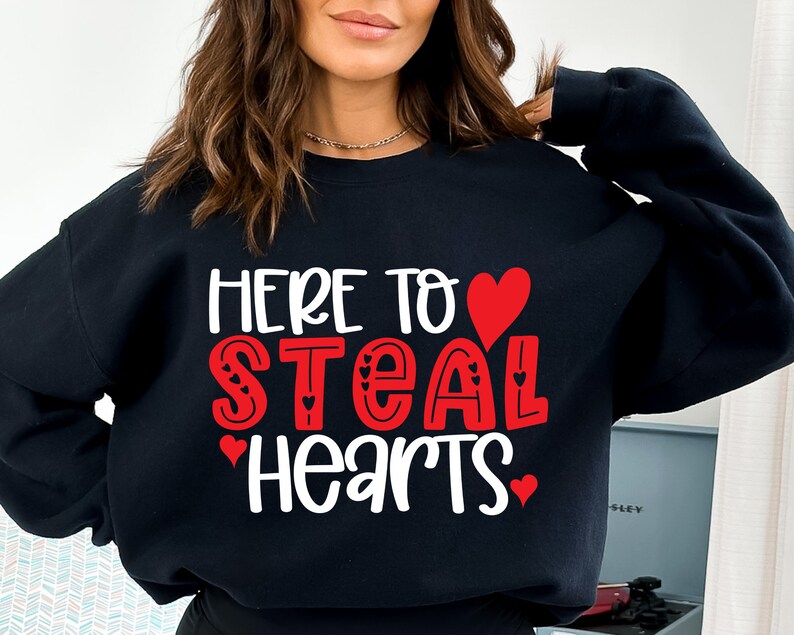 May include: Black sweatshirt with the text "HERE TO STEAL HEARTS" in white and red lettering. The word "STEAL" is in red with heart accents. A red heart is above the word "STEAL".