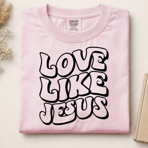 Pu&ograve; includere: Una t-shirt a maniche corte rosa chiaro con il testo nero LOVE LIKE JESUS in un carattere retro audace e ondulato. Il capo presenta un girocollo e un'etichetta del marchio Comfort Colors stampata all'interno della zona del colletto.
