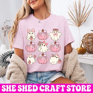 K&ouml;nnte beinhalten: Ein hellrosa T-Shirt mit einem Rastermuster aus neun Illustrationen. Das Design zeigt wei&szlig;e Geister mit rosa Schleifen und Blumen sowie rosa K&uuml;rbisse, die mit Schleifen verziert sind und in drei Reihen zu je drei Motiven angeordnet sind.