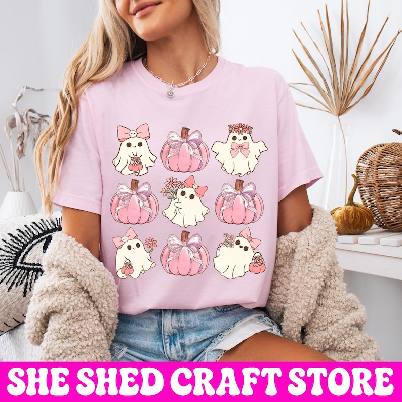 K&ouml;nnte beinhalten: Ein hellrosa T-Shirt mit einem Rastermuster aus neun Illustrationen. Das Design zeigt wei&szlig;e Geister mit rosa Schleifen und Blumen sowie rosa K&uuml;rbisse, die mit Schleifen verziert sind und in drei Reihen zu je drei Motiven angeordnet sind.