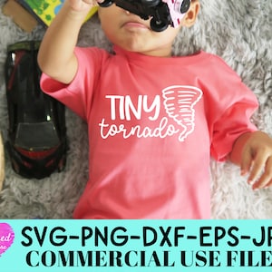 May include: A coral-colored short-sleeve t-shirt featuring the white text "TINY tornado" and a graphic illustration of a tornado. The shirt is worn by a child who is holding a small toy vehicle, with other toys visible in the surrounding area.