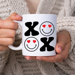 Pu&ograve; includere: Tazza in ceramica bianca con un design in bianco e nero che presenta le lettere "XO" e due facce sorridenti con cuori rossi come occhi.