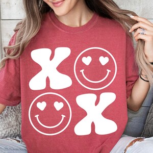 Pu&ograve; includere: Maglietta rossa con un design grafico bianco di due facce sorridenti con cuori per gli occhi e le lettere "XO" in stile retr&ograve;.