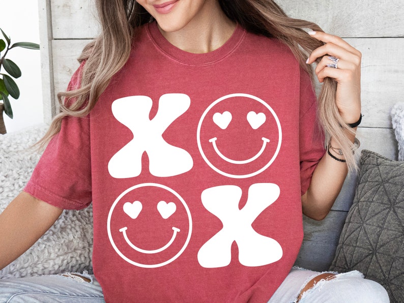 Pu&ograve; includere: Maglietta rossa con un design grafico bianco di due facce sorridenti con cuori per gli occhi e le lettere "XO" in stile retr&ograve;.