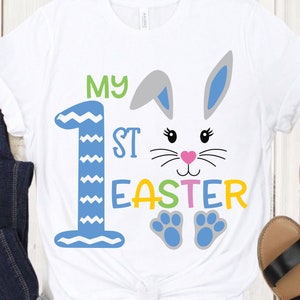 May include: White t-shirt with a blue and white number one, the words "My 1st Easter" in green, pink, yellow, and blue, and a bunny face with blue ears, pink nose, and gray whiskers.