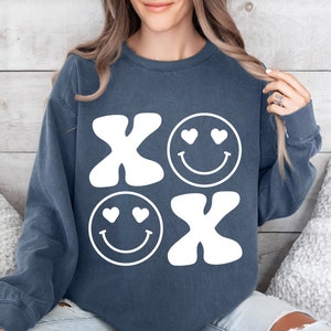 Pu&ograve; includere: Una felpa blu scuro con un design grafico bianco di due facce sorridenti con cuori per gli occhi e le lettere "XO" in stile retr&ograve;.