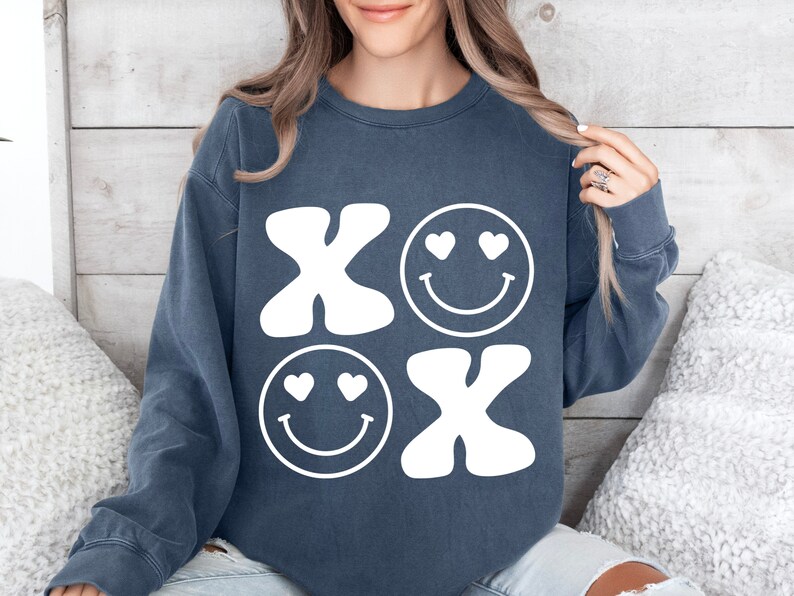 Pu&ograve; includere: Una felpa blu scuro con un design grafico bianco di due facce sorridenti con cuori per gli occhi e le lettere "XO" in stile retr&ograve;.
