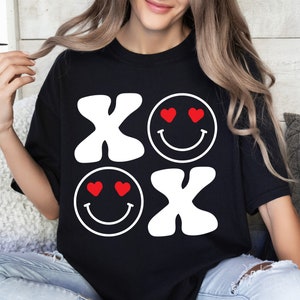 Pu&ograve; includere: Maglietta nera con un design grafico bianco delle lettere "XO" e due faccine sorridenti con cuori per gli occhi. Le faccine sorridenti sono rosse.