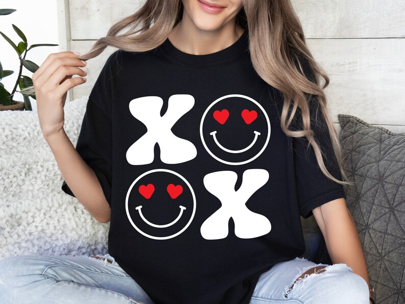 Pu&ograve; includere: Maglietta nera con un design grafico bianco delle lettere "XO" e due faccine sorridenti con cuori per gli occhi. Le faccine sorridenti sono rosse.