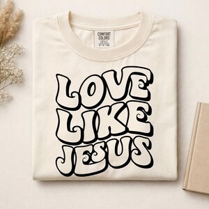 Pu&ograve; includere: Una t-shirt color crema con il testo nero LOVE LIKE JESUS in un carattere audace, ondulato e di ispirazione retr&ograve;. La maglia presenta un girocollo e un'etichetta del marchio Comfort Colors stampata all'interno del colletto.