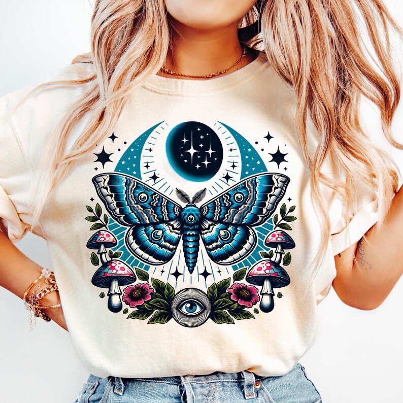 May include: A white t-shirt with a blue and black moth design. The moth is surrounded by a crescent moon, stars, mushrooms, and flowers. There is also a blue eye in the center of the design.
