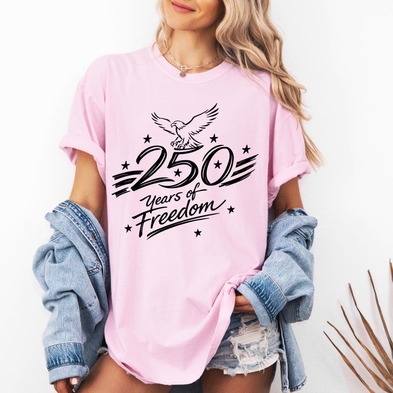 K&ouml;nnte beinhalten: Ein hellrosa T-Shirt mit einer schwarzen Grafik eines Adlers &uuml;ber dem Text 250 Years of Freedom, umgeben von kleinen Sternen. Das T-Shirt hat kurze &Auml;rmel und einen Rundhalsausschnitt und wird mit einer hellblauen Jeansjacke und Jeansshorts getragen.