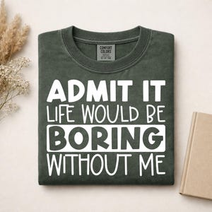 May include: A dark olive green crew neck t-shirt featuring the white printed text ADMIT IT LIFE WOULD BE BORING WITHOUT ME. The shirt has a Comfort Colors brand label on the inside collar and is folded neatly on a flat surface.