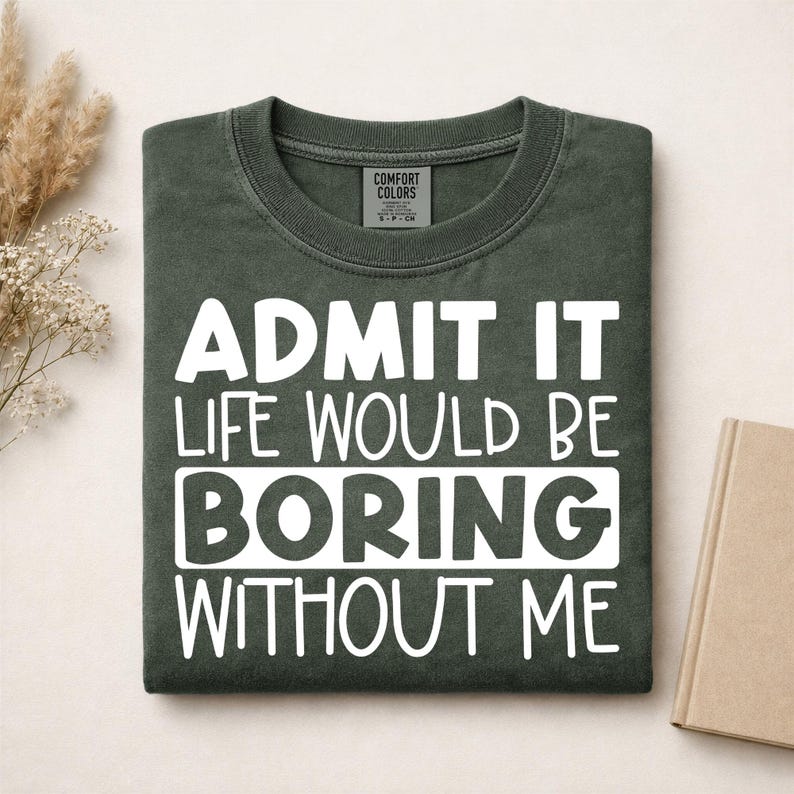 May include: A dark olive green crew neck t-shirt featuring the white printed text ADMIT IT LIFE WOULD BE BORING WITHOUT ME. The shirt has a Comfort Colors brand label on the inside collar and is folded neatly on a flat surface.