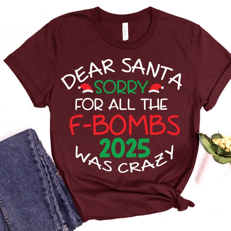 May include: Burgundy t-shirt with the text "DEAR SANTA, SORRY FOR ALL THE F-BOMBS 2025 WAS CRAZY" in white, green, and red. The text includes Santa hats. The shirt is paired with blue jeans.