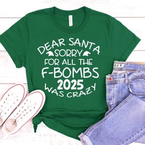 May include: A green t-shirt with the text "DEAR SANTA SORRY FOR ALL THE F-BOMBS 2025 WAS CRAZY" in white. The shirt is paired with blue jeans and white sneakers.