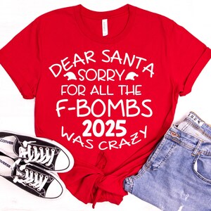 May include: A red t-shirt with the text "DEAR SANTA SORRY FOR ALL THE F-BOMBS 2025 WAS CRAZY" in white. The shirt is paired with blue jeans and black Converse sneakers.