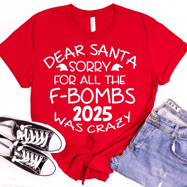 May include: A red t-shirt with the text "DEAR SANTA SORRY FOR ALL THE F-BOMBS 2025 WAS CRAZY" in white. The shirt is paired with blue jeans and black Converse sneakers.