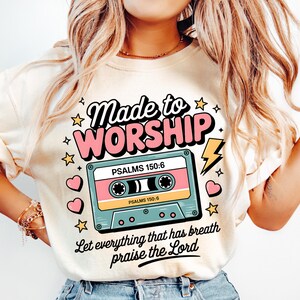 May include: A cream-colored t-shirt with a retro-style design featuring a cassette tape and the text "Made to Worship" with the Bible verse "Psalms 150:6" and the phrase "Let everything that has breath praise the Lord".