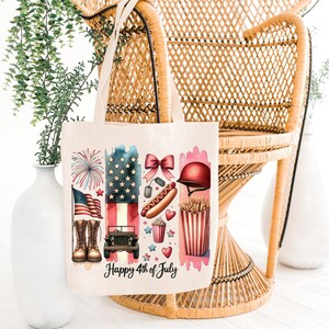 K&ouml;nnte beinhalten: Eine wei&szlig;e Canvas-Tragetasche mit einem rot-wei&szlig;-blauen Design mit Feuerwerk, einem Jeep, einem Hotdog, Popcorn und einem Helm. Der Text "Happy 4th of July" ist auf die Tasche gedruckt.