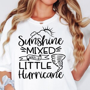 May include: White t-shirt with the text "Sunshine Mixed with a Little Hurricane" in black script. The design includes a sun, clouds, and a tornado graphic. The shirt is a casual, comfortable style.