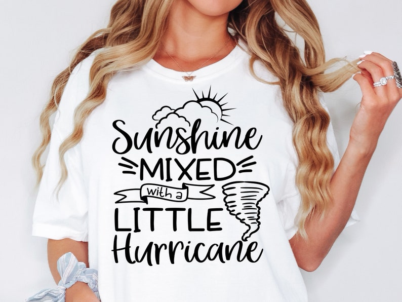 May include: White t-shirt with the text "Sunshine Mixed with a Little Hurricane" in black script. The design includes a sun, clouds, and a tornado graphic. The shirt is a casual, comfortable style.