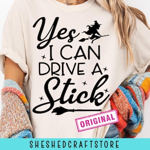 May include: Cream-colored t-shirt with the phrase "Yes I Can Drive A Stick" in black script and block letters, with a witch on a broom graphic. The word "ORIGINAL" is in a pink rectangle.