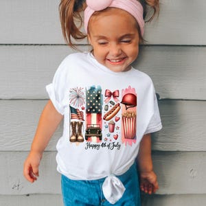 K&ouml;nnte beinhalten: Wei&szlig;es T-Shirt mit einem bunten Grafikdesign, das eine amerikanische Flagge, Feuerwerk, einen Jeep, einen Hotdog, Popcorn und einen roten Helm zeigt. Der Text "Happy 4th of July" ist auf das Shirt gedruckt.
