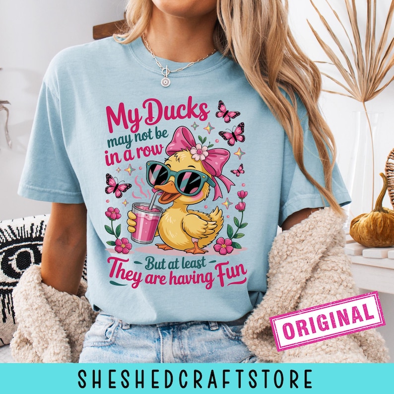 K&ouml;nnte beinhalten: Ein hellblaues T-Shirt mit einer Grafik eines gelben Entchens, das eine Sonnenbrille und eine rosa Schleife tr&auml;gt und ein rosa Getr&auml;nk h&auml;lt. Der Text lautet: My Ducks may not be in a row But at least They are having Fun. Rosa Schmetterlinge und Blumen.