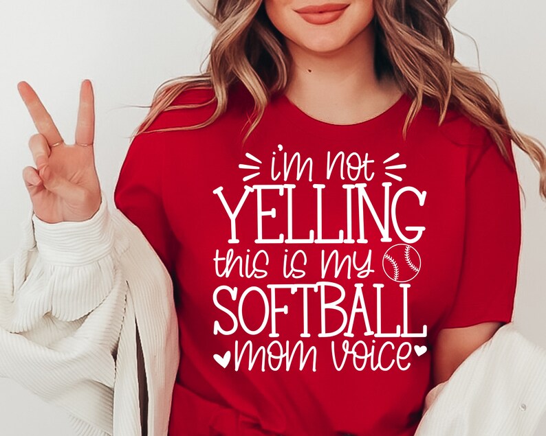 May include: Red t-shirt with white text that reads "I'm not yelling this is my softball mom voice" with a white baseball graphic.