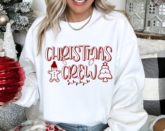 May include: A white crewneck sweatshirt featuring the red text CHRISTMAS CREW. The design includes a gingerbread person wearing a hat, a string of lights, and a stylized Christmas tree. The lettering is in a bold, rounded font style.