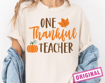 May include: Cream-colored t-shirt with the text "ONE Thankful TEACHER" in brown and orange. A pumpkin and a maple leaf graphic are included in the design. The word "ORIGINAL" is printed on a pink rectangle.