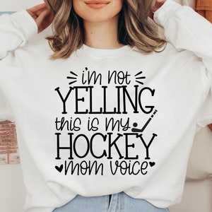 May include: White sweatshirt with black text that reads "I'm not yelling this is my hockey mom voice" with a hockey stick and puck graphic.