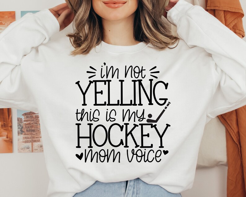May include: White sweatshirt with black text that reads "I'm not yelling this is my hockey mom voice" with a hockey stick and puck graphic.