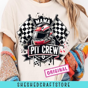 Może przedstawiać: Beżowa koszulka z graficznym wzorem przedstawiającym kask wyścigowy, szachownice i napisem "MAMA PIT CREW". Projekt zawiera tekst "ORIGINAL" i "SHESHEDCRAFTSTORE."