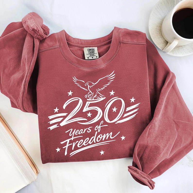 K&ouml;nnte beinhalten: Ein ziegelrotes Sweatshirt mit Rundhalsausschnitt und einem wei&szlig;en Grafikdruck eines Wei&szlig;kopfseeadlers &uuml;ber der Zahl 250 sowie dem Text Years of Freedom, akzentuiert durch kleine Sterne und horizontale Linien. Mit Comfort Colors Etikett.
