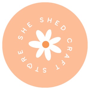 K&ouml;nnte beinhalten: Ein pfirsichfarbener Kreis mit dem Text "She Shed Crafts" in wei&szlig;en Buchstaben um eine wei&szlig;e Margerite mit gelber Mitte.