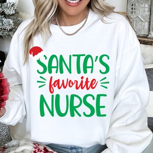 以下が含まれることがあります： 前面に緑と赤の文字で「SANTA'S favorite NURSE」とプリントされた白い長袖スウェットシャツ。デザインには、「SANTA'S」という単語の上に、赤と白のサンタクロースの帽子の小さなグラフィックが施されており、祝祭的なスタイルに仕上がっています。